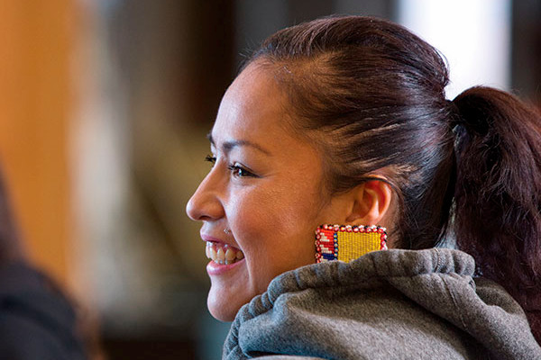 An Indigenous woman wearing colourful earrings smiles at others off-camera. She's sitting in Chinook Lodge Resource Centre.